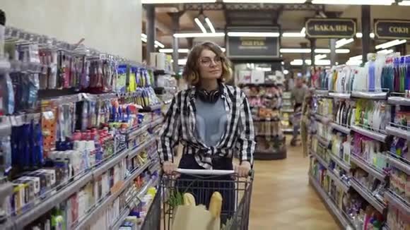 现代女孩戴着眼镜戴着耳机脖子上戴着购物车走在超市里四处闲逛视频的预览图
