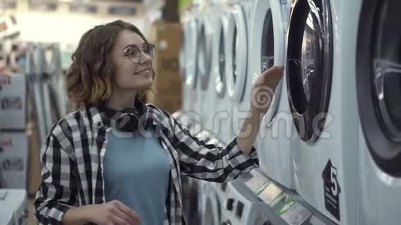 一个年轻的积极的女人穿着格子衬衫在家用电器商店里选择洗衣机打开门然后视频的预览图