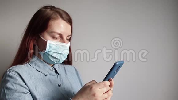 一个戴着医用口罩的年轻女孩正在智能手机上对着灰色的墙壁说话视频的预览图