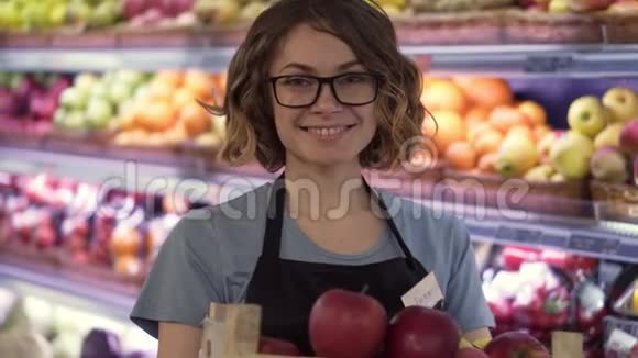 美丽的微笑年轻的女超市员工穿着黑色围裙手里拿着一盒装满苹果的架子视频的预览图