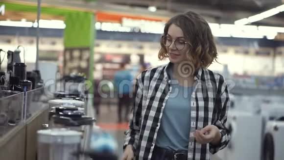 在电器商店里一位穿着格子衬衫的黑发卷发女士通过观看和拿着来选择搅拌机购物视频的预览图