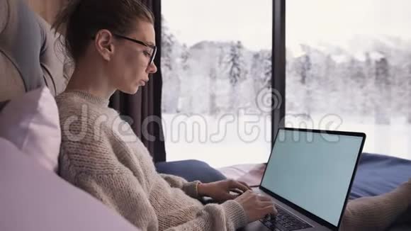 一幅有魅力的自由职业者的画像她戴着眼镜穿着一件毛衣穿着长袜坐在生态屋的床上视频的预览图