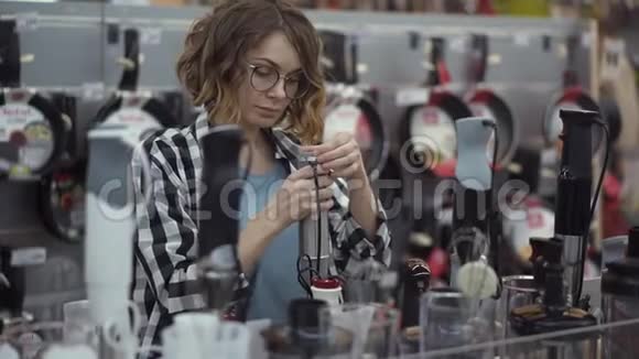 在电器商店里一个穿着格子衬衫的漂亮卷发女人通过观看和拿着搅拌器棒视频的预览图