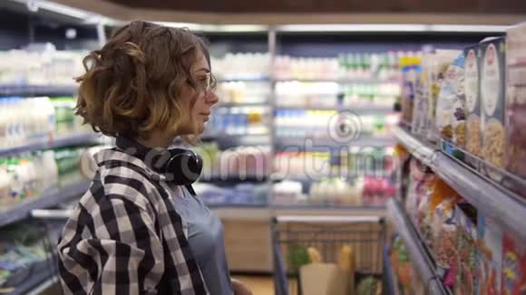 一位戴着耳机的年轻女士在杂货店里选择燕麦片和薄片这是一种完美的食物视频的预览图
