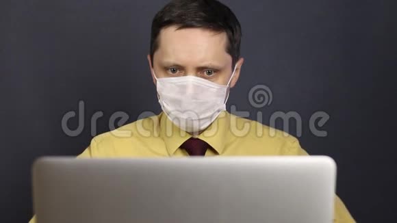 一个戴着医用口罩的男人用笔记本电脑通过互联网阅读新闻困惑地打手势抓着他的下巴他摇了摇他的视频的预览图