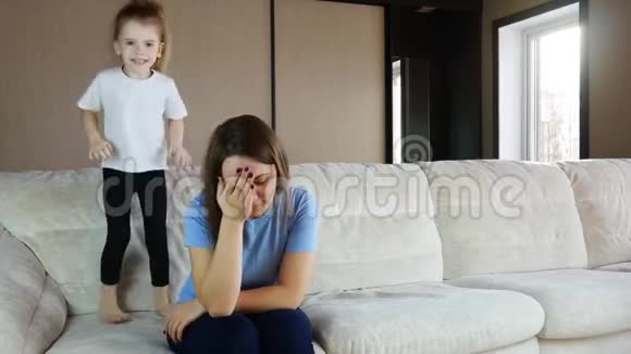 母亲坐在沙发上沉思活泼的孩子女儿在跳跃玩耍视频的预览图