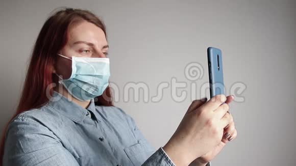 一个戴着医用口罩的年轻女孩正在智能手机上对着灰色的墙壁说话冠状病毒病毒概念视频的预览图