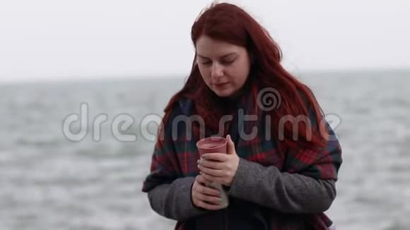 小女孩拿着一个金属热水瓶在靠近大海的河堤上放着一杯热饮料外面大风头发长在视频的预览图