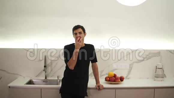 年轻人在家里厨房里吃红苹果穿着黑色衣服的家伙吃着新鲜苹果看着视频的预览图