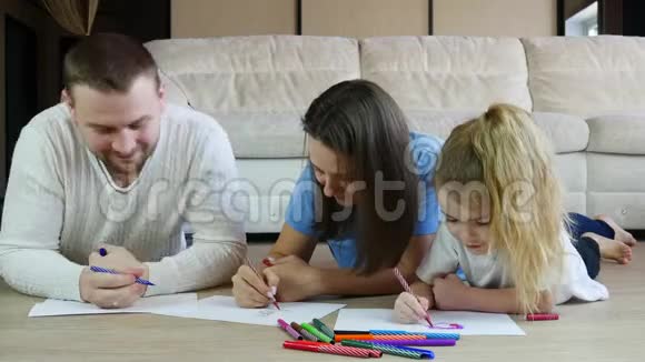 年轻的父母画着彩色图画可爱的女儿躺在温暖的地板上视频的预览图