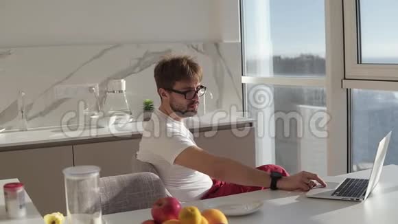 戴着眼镜的胡子男戴着笔记本电脑坐在家里的厨房里厨房里开着全景窗年轻妻子视频的预览图