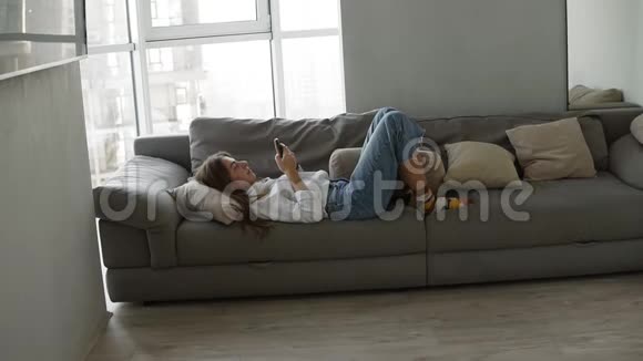在客厅里疲惫的母亲躺在沙发上戴着智能手机可爱的女儿和父亲突然从后面出现了视频的预览图