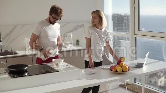 美丽的白种人年轻男子在家里的厨房为两人准备早餐在碗里搅拌鸡蛋妻子过来视频的预览图