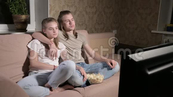 年轻可爱的夫妇坐在客厅里拥抱看电视吃爆米花视频的预览图