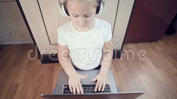 一个7岁的聪明的小女孩戴着白色耳机手里拿着一台笔记本电脑正在她的地板上推着视频的预览图