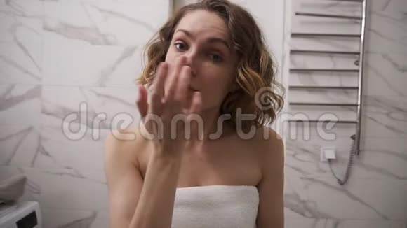 一位身穿白色大毛巾的年轻女士正在眼部下面的面部涂奶油按摩动作看镜子视频的预览图