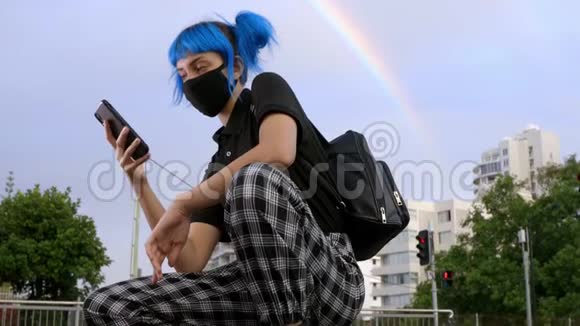年轻女性戴着时尚的医用口罩蓝色头发看着智能手机视频的预览图
