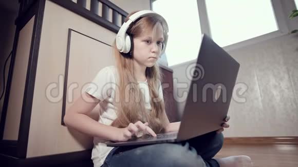 一个7岁的聪明的小女孩戴着白色耳机手里拿着一台笔记本电脑正在她的地板上推着视频的预览图