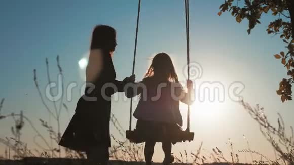 幸福的年轻母亲和小女儿在阳光下荡秋千的剪影坐在木秋千上的漂亮女孩视频的预览图
