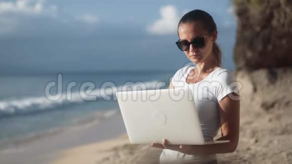 戴墨镜的女性自由职业者手提电脑类型和工作在热带海滩视频的预览图