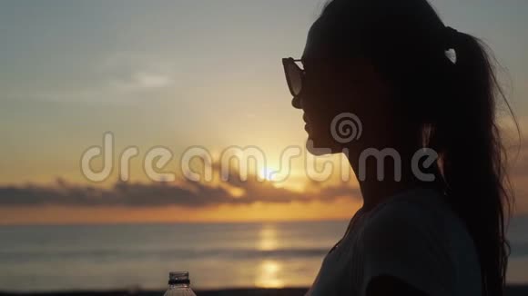 戴太阳镜的女人在日落时从瓶子里喝清水视频的预览图