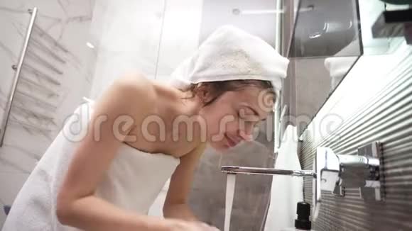 一个女人站在洗手池附近头上戴着白色毛巾身体用自来水冲洗她的脸视频的预览图