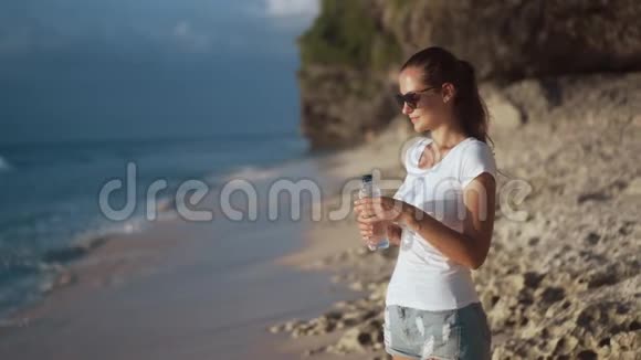 戴太阳镜的漂亮年轻女子从沙滩上的瓶子里喝着清澈的水视频的预览图