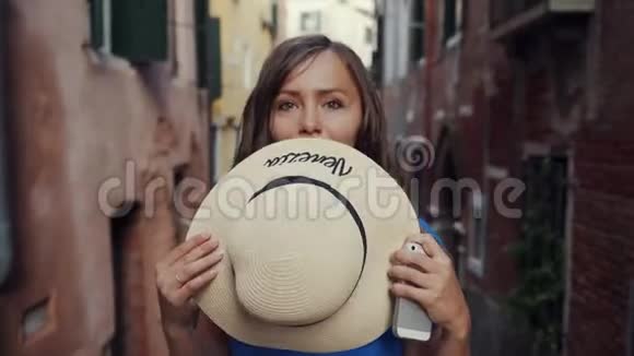 布鲁内特姑娘站在威尼斯狭窄的街道上用草帽遮住她的脸视频的预览图