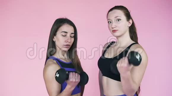 两个年轻的运动女孩在粉红色的背景上用哑铃握手健身和运动慢动作视频的预览图