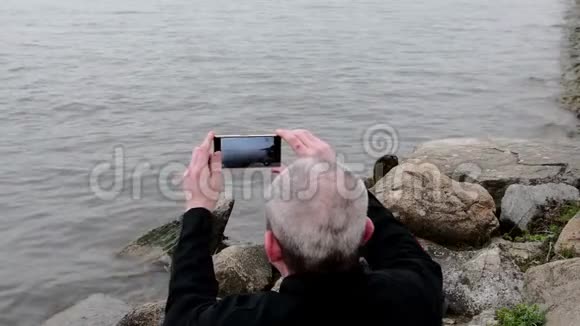 坐在湖岸上的中年老人孤独的成熟男人用手机在银行录制视频视频的预览图