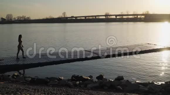 年轻的运动女孩在美丽的日落背景下沿着湖下的浮舟慢动作对Steadicam的射击视频的预览图