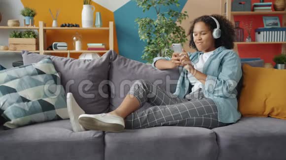 快乐的非裔美国女士使用智能手机在家中享受耳机中的音乐视频的预览图