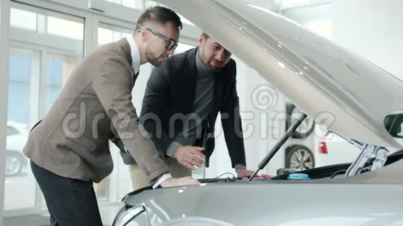 性格开朗的男性销售经理和客户在室内引擎盖下观看汽车引擎视频的预览图
