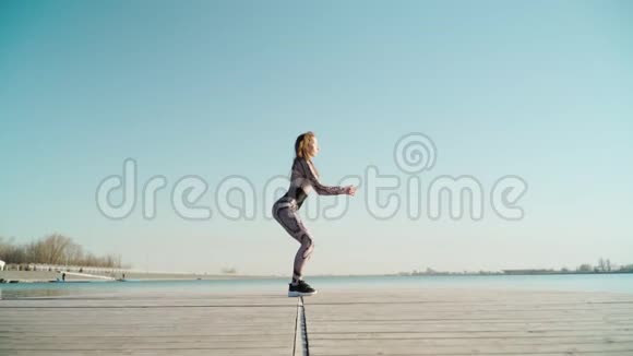 年轻健康的黑发美女穿着紧身裤黑色运动鞋在蓝湖边做运动视频的预览图