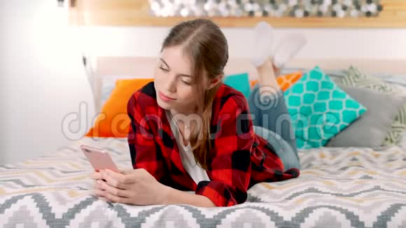 一个年轻漂亮的白人女人躺在床上微笑着手里拿着一部手机和她的朋友发短信视频的预览图