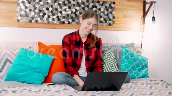 一个年轻可爱的白人女人正坐在床上两条腿交叉着在现代卧室里微笑着用笔记本电脑进行视频通话视频的预览图