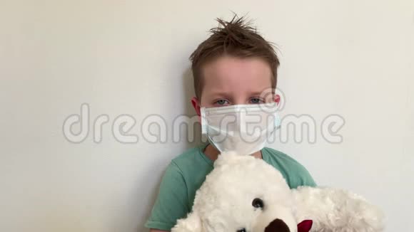 一个带防护面罩的孩子和一只泰迪熊坐在家里在隔离看着相机眨着眼睛视频的预览图