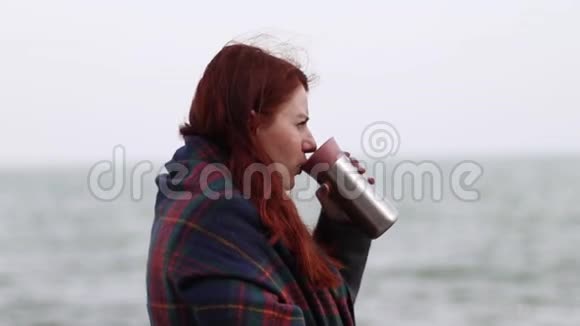 年轻的红头发女孩拿着一个金属热水瓶在靠近大海的河堤上放着一杯热饮料视频的预览图