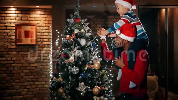 一个孩子正在用新年玩具装饰一棵圣诞树坐在他父亲肩膀上视频的预览图