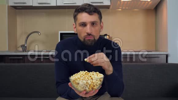 男人坐在沙发上吃透明玻璃盘子里的爆米花看电视上的娱乐节目视频的预览图