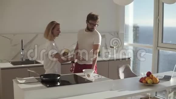 年轻迷人的一对夫妇穿着家庭服装在明亮的钉厨房烹饪简单的早餐快乐的妻子吻她视频的预览图