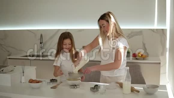 母亲在现代舒适的白色厨房里帮助和教她可爱的女儿做蛋糕或煎饼妈妈添加面粉视频的预览图
