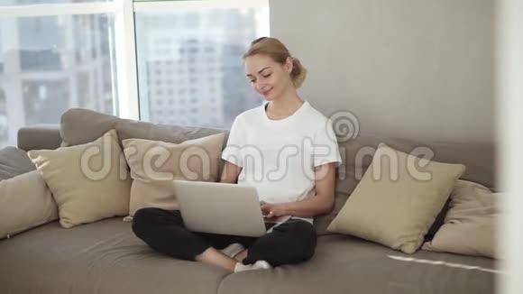 微笑的女性自由职业者坐在沙发上穿着家庭服装自由职业者从家里在笔记本电脑上输入电子邮件重点视频的预览图