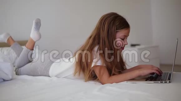 长发欧洲女孩在笔记本电脑上打字微笑的小女孩带着兴趣看着笔记本电脑躺在床上侧视图视频的预览图