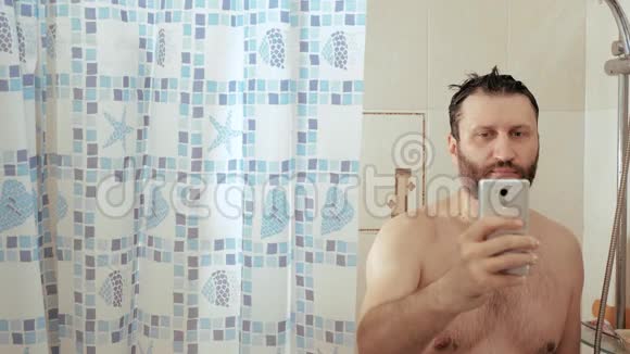 淋浴的胡子男在智能手机中使用互联网阅读新闻社交网络他的头上涌着水慢慢视频的预览图