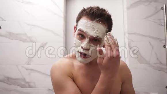 年轻帅哥的肖像正在涂上新鲜的白色厚厚的面具并在脸上擦洗天然有机化妆品视频的预览图