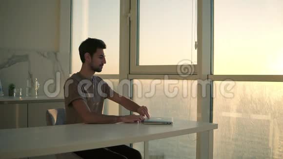 欧洲黑头发男人自由职业者打开笔记本电脑工作坐在演播室全景窗口附近的年轻人视频的预览图