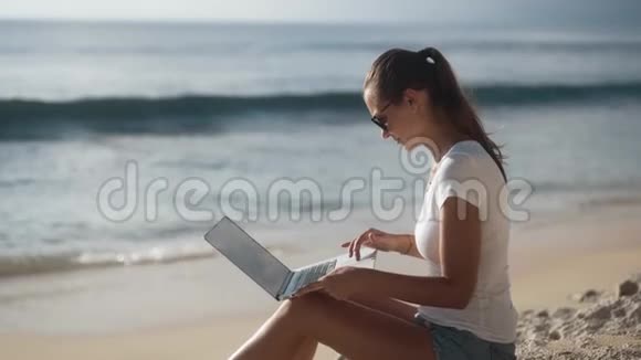 戴太阳镜的女性自由职业者使用笔记本电脑在海滩上工作背景是大海视频的预览图