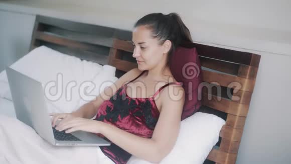 美丽开朗的女孩在笔记本电脑上工作在床上喝橙汁视频的预览图