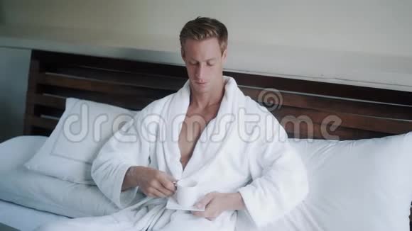 穿浴袍的年轻人在笔记本电脑附近的床上喝咖啡视频的预览图